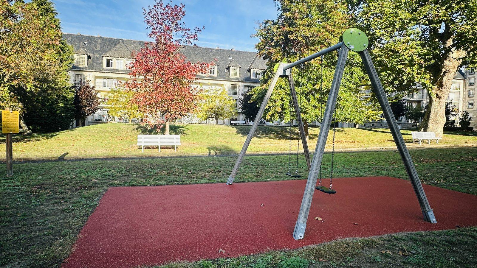 Le portique à balançoires sur son sol souple rouge, devant la pelouse et l'immeuble de la résidence.