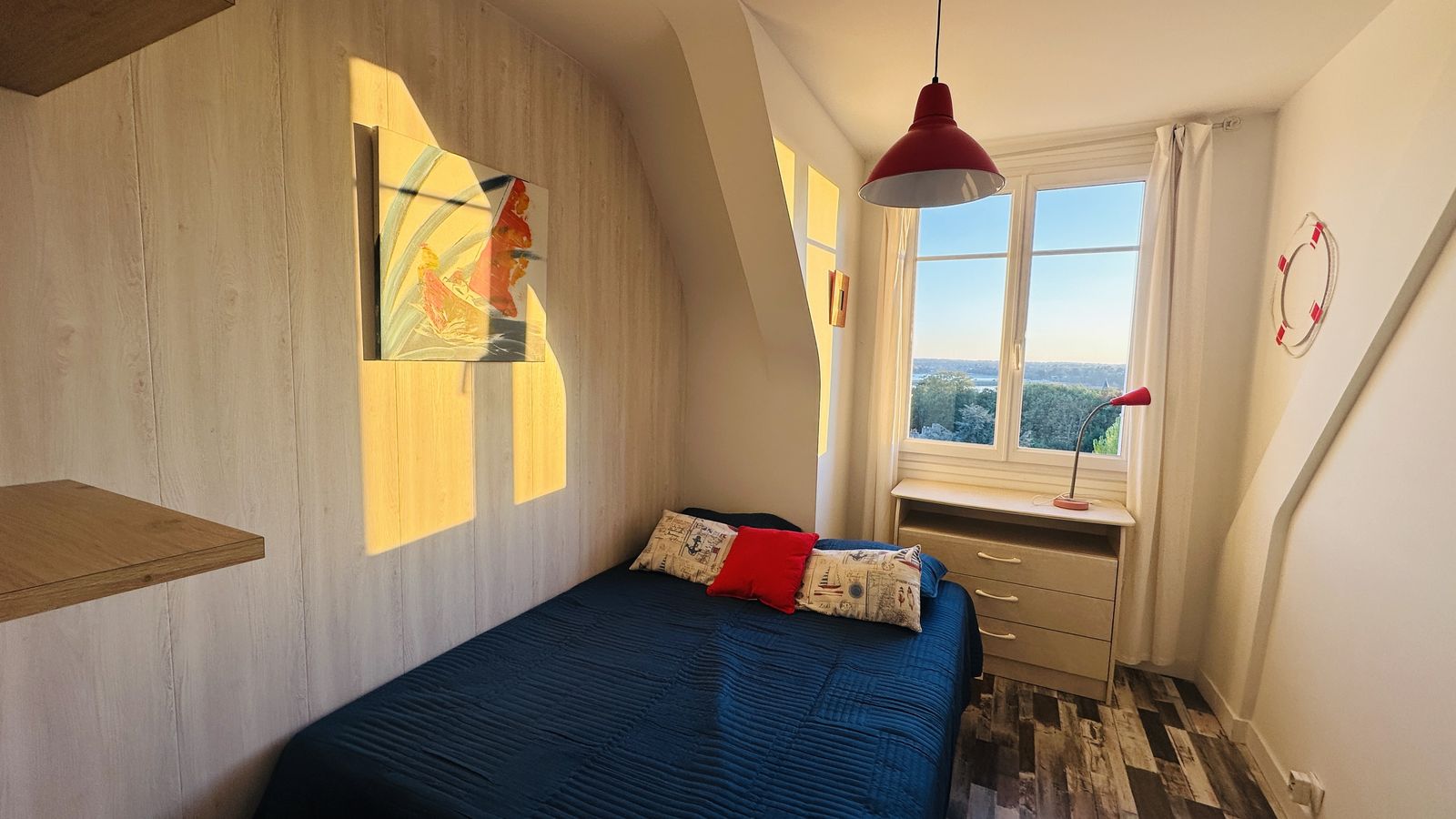 Chambre mansardée : un lit double au couvre-lit bleu marine, garni de coussins, une suspension rouge, une commode sous la fenêtre par laquelle on aperçoit la mer, et une petite lampe rouge. Au mur, une toile de voilier dans le soleil.