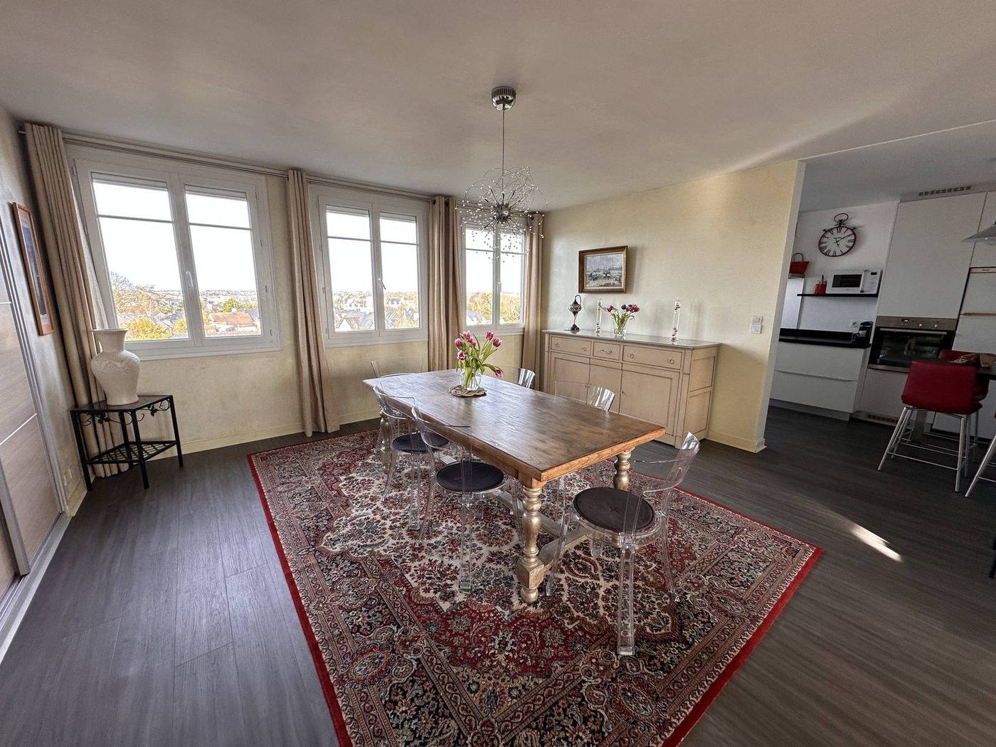 Coin repas : grande table en bois et six chaises transparentes sur un tapis rouge à motifs, devant trois fenêtres qui donnent sur les toits d'ardoise ; un buffet, un bouquet de tulipes, et à droite l'ouverture sur la cuisine.