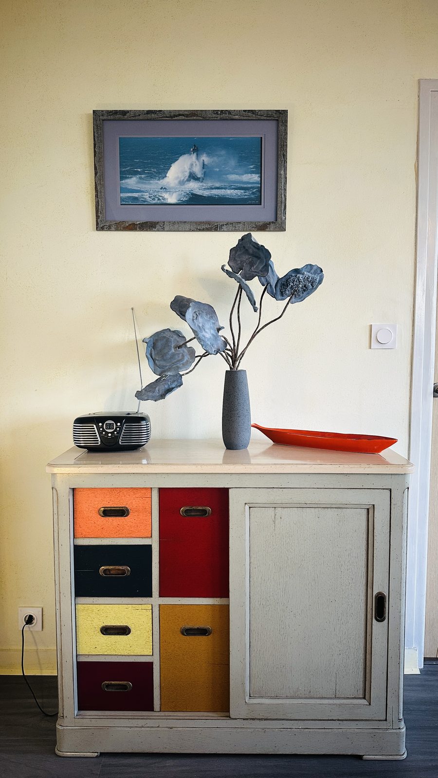 Dans l'entrée, une commode aux tiroirs peints en orange, bleu, jaune et rouge, surmontée d'un bouquet de fleurs séchées dans un vase ; au mur, une marine encadrée.